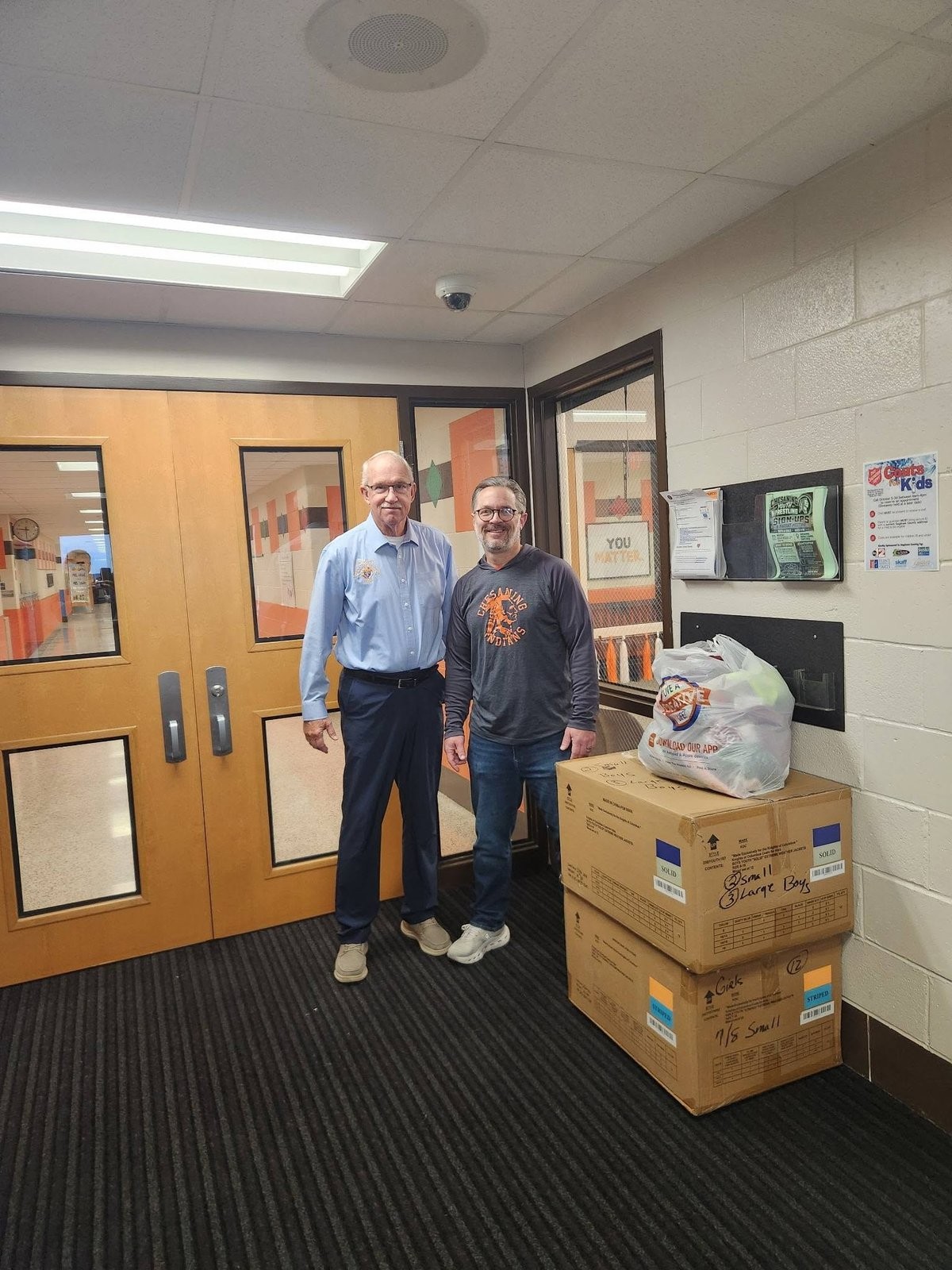 Two men with boxes and a bag of donated coats inside a school entrance