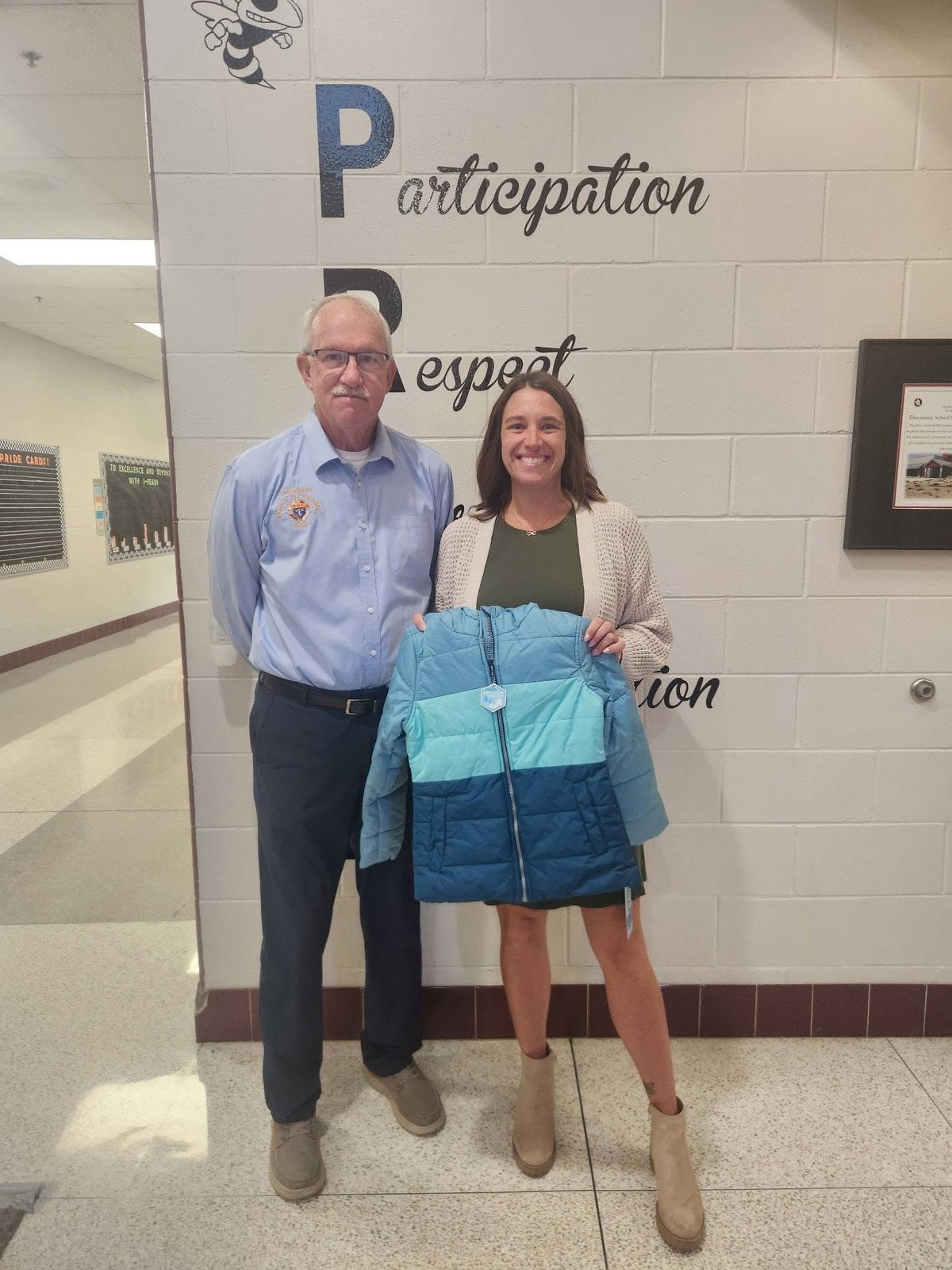 A man and a woman holding a donated teal winter coat in a school hallway